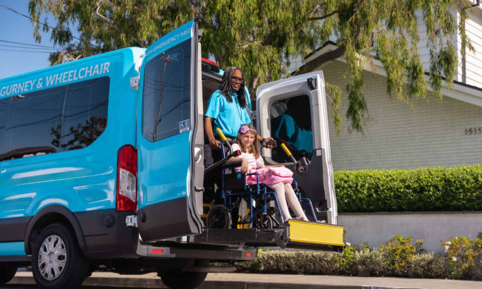 Male HopSkipDrive in a blue shirt uses a wheelchair lift to help a disabled student out of a van.
