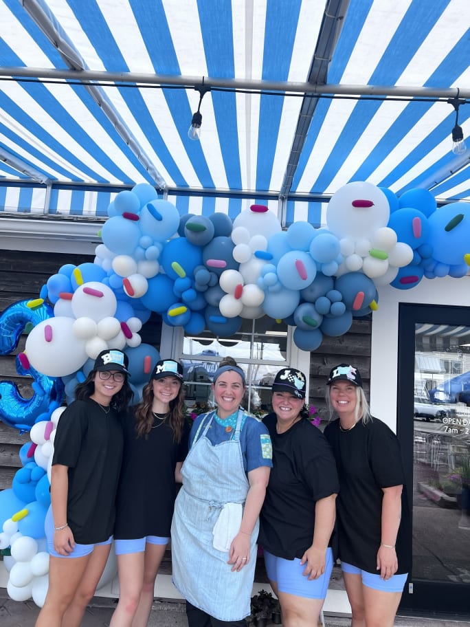 Lillian James team members stand with a baker in front of an arch of blue balloons.