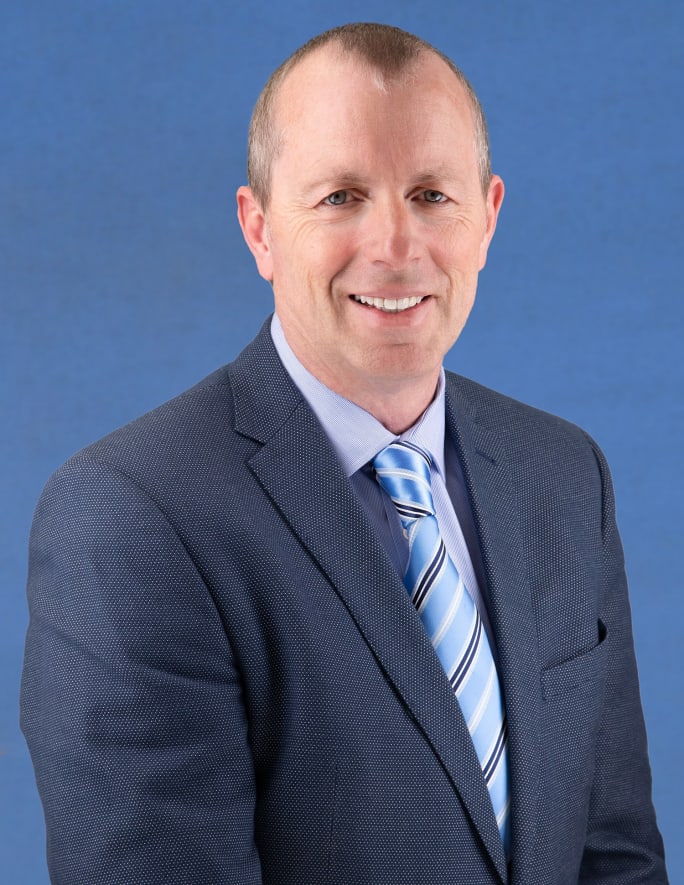 A business portrait of Ben Carfrae in a blue suit and tie in front of a blue background.