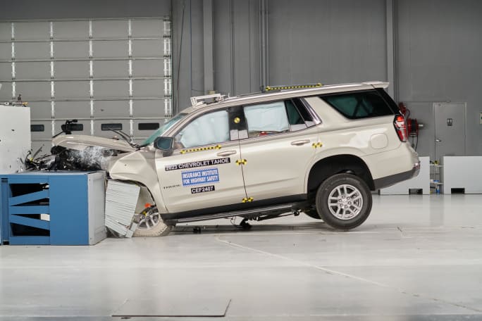 An off-white Chevrolet Tahoe is shown with major damage to its front end and its air bags deployed in a crash testing facility.