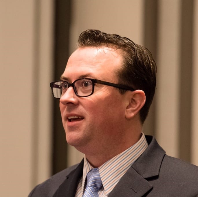 A portrait of a man in a suit and glasses looking off camera left.