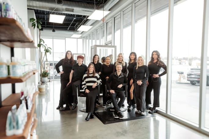 Dressed in black, a group of stylists pose in the salon between retail shelves and big storefront windows.