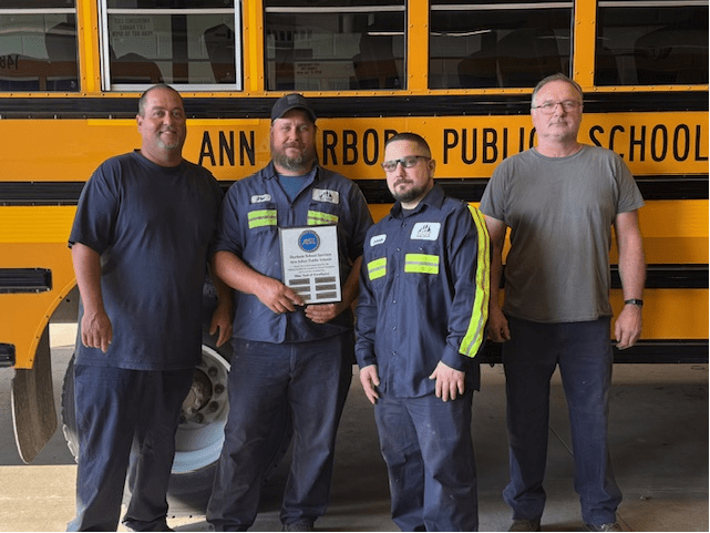 Four Durham School Services technicians stand in front of a yellow school bus inside a garage