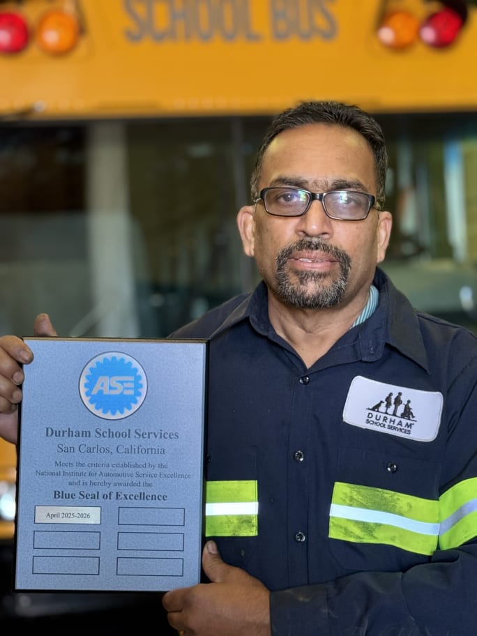 a man poses with an ASE plaque with a school bus in the background