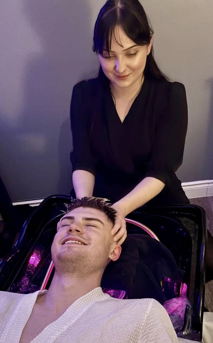 A male client smiles as he relaxes and an esthetician gives him a scalp treatment.