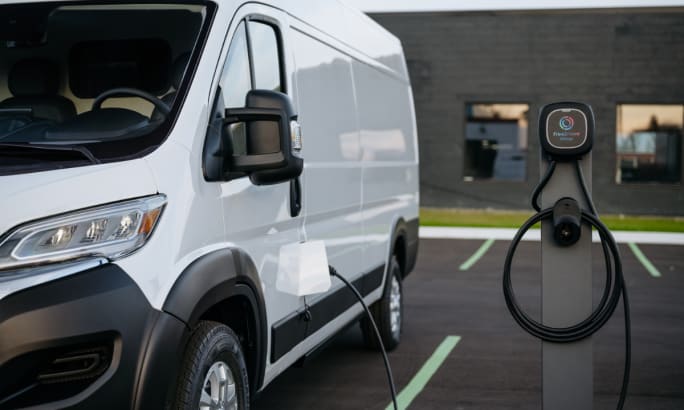Front left side of a 2025 Ram ProMaster EV with charging port open and connected to an EV charger.