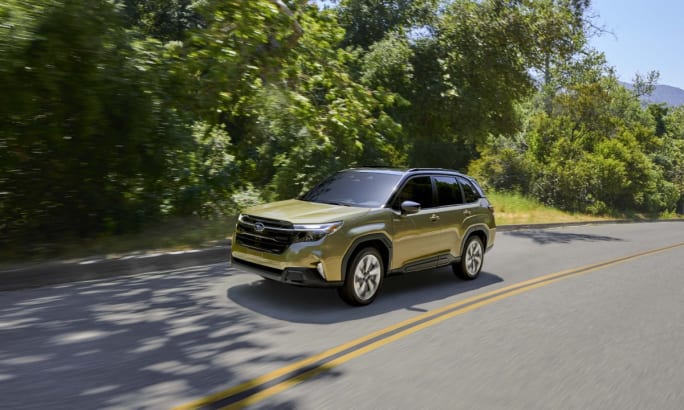 A green 2025 Subaru Forester Hybrid driving on a road with foliage in the background.