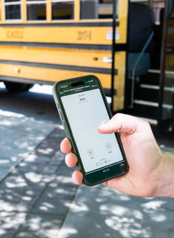 Hand holds a smartphone displaying a digital vehicle inspection checklist in front of a school bus.