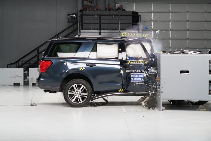 A dark blue Ford Expedition is shown with its front end entirely crumpled and air bags deployed in a crash testing facility.