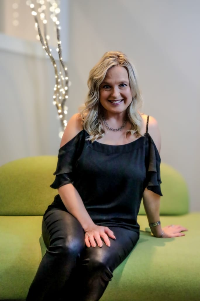 Amy Mayfield Lusche seated on the edge of a lime green sofa with twinkling lights behind her.