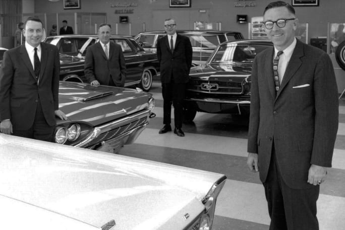 A black and white photo shows men in suits standing around a Ford dealership showroom.