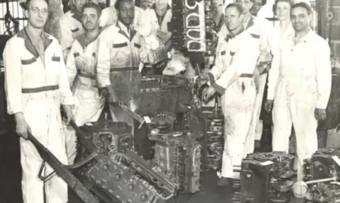 A black and white photo shows employees posing with vehicle transmissions.