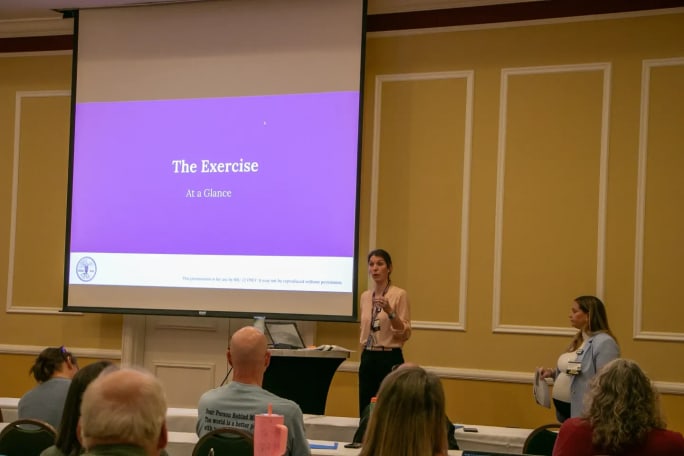 Two presenters speak in front of a screen displaying “The Exercise” during a school safety training session with attendees seated and listening.
