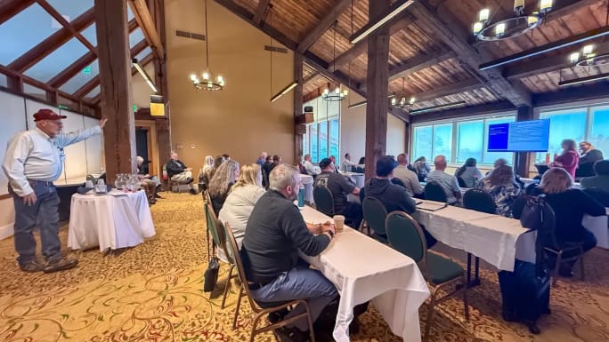 Audience seated at tables listens to a presentation during a school safety conference, with a speaker presenting slides at the front of the room.