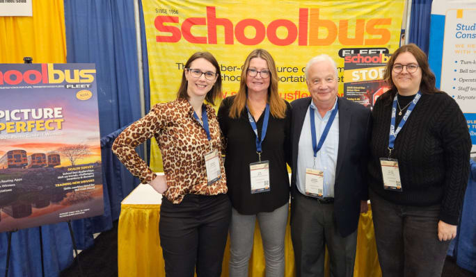 four people stand together in a trade show booth with a school bus fleet sign in the background