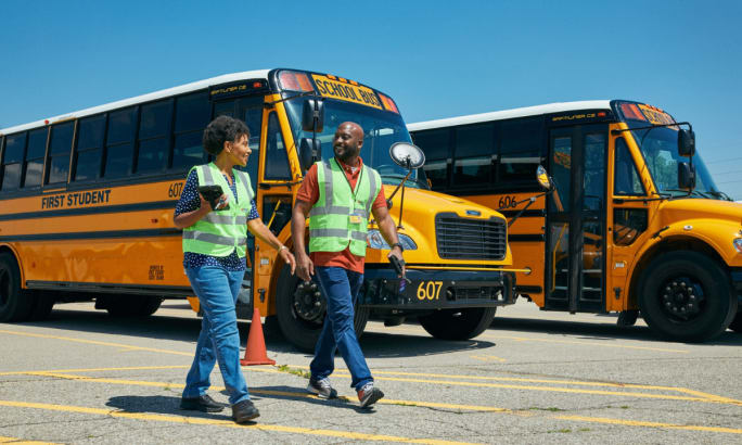 Two people in high visibility vest walking in front of parked school buses.