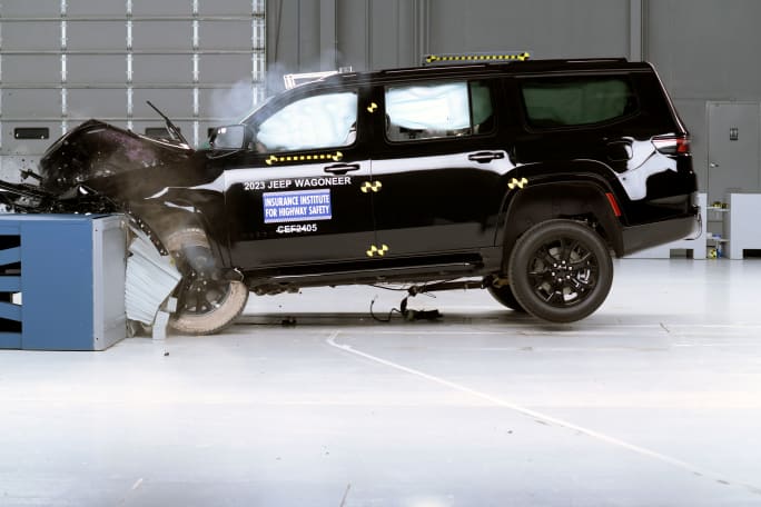 A black Jeep Wagoneer is shown with its front bumper heavily damaged and air bags deployed in a crash testing facility.