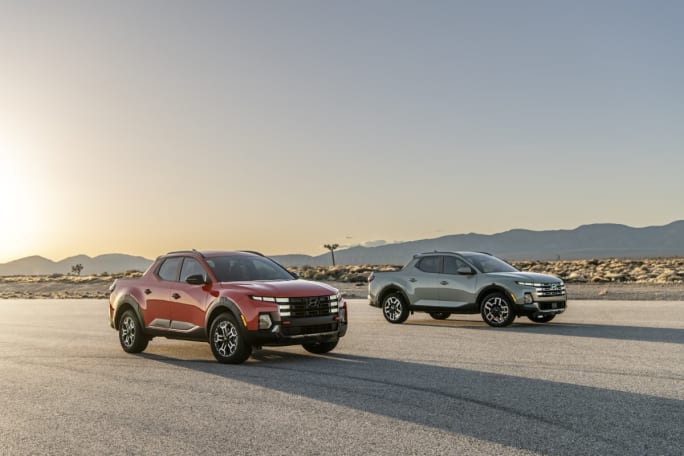 Two 2025 Hyundai Santa Cruz trucks, one red and one pale green, parked on a road with the sunset and mountains in background.