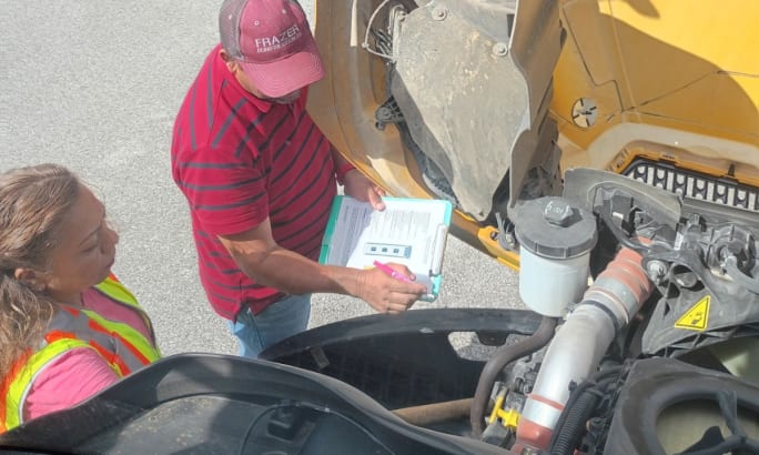 A man with a clipboard shows a school bus driver what to look for during an inspection.