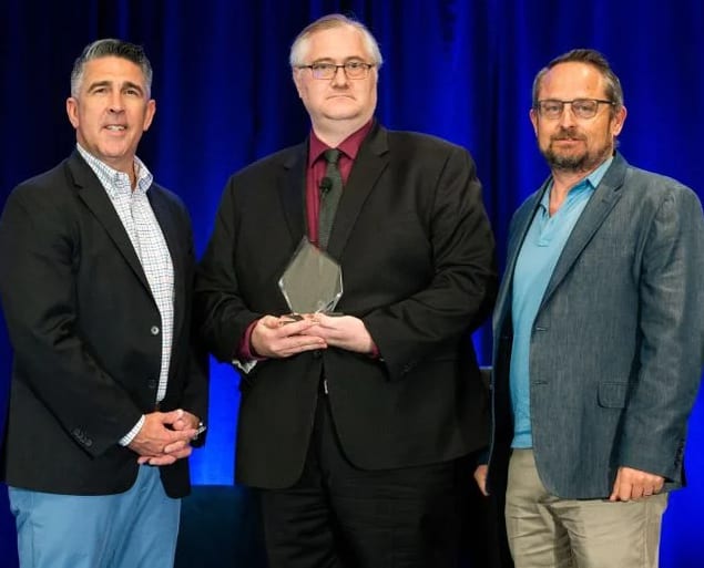 Three men stand in front of blue curtains for an award photo. 