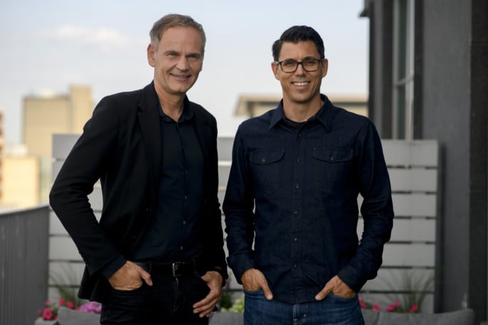 Two men pose in on an outdoor balcony in dark colored business casual attire. 