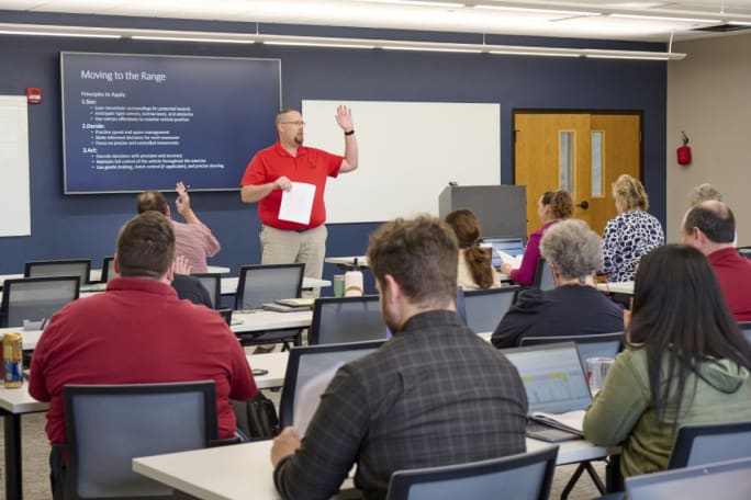 Adult classroom setting with a teacher standing in front of a projector screen addressing the class.