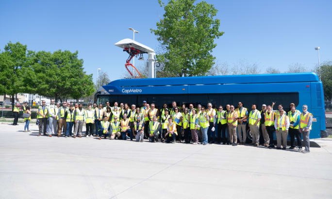 Employees pose in front of a blue CapMetro automated bus at an overhead charging station.
