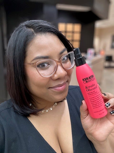 A woman with black hair, large eyeglass frames, holding a hair product.