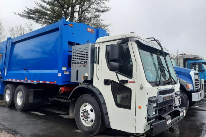 A white Class 8 Mack LR truck with a blue refuse body is shown at a media event celebrating the vehicle's delivery.