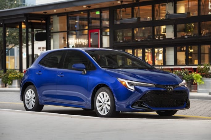 A blue 2025 Toyota Corolla Hatchback parked in front of city building.