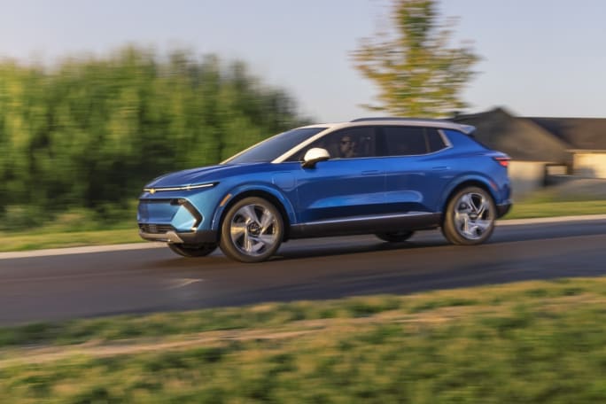 Blue Chevrolet Equinox EV SUV driving on a road with a house and greenery in background.