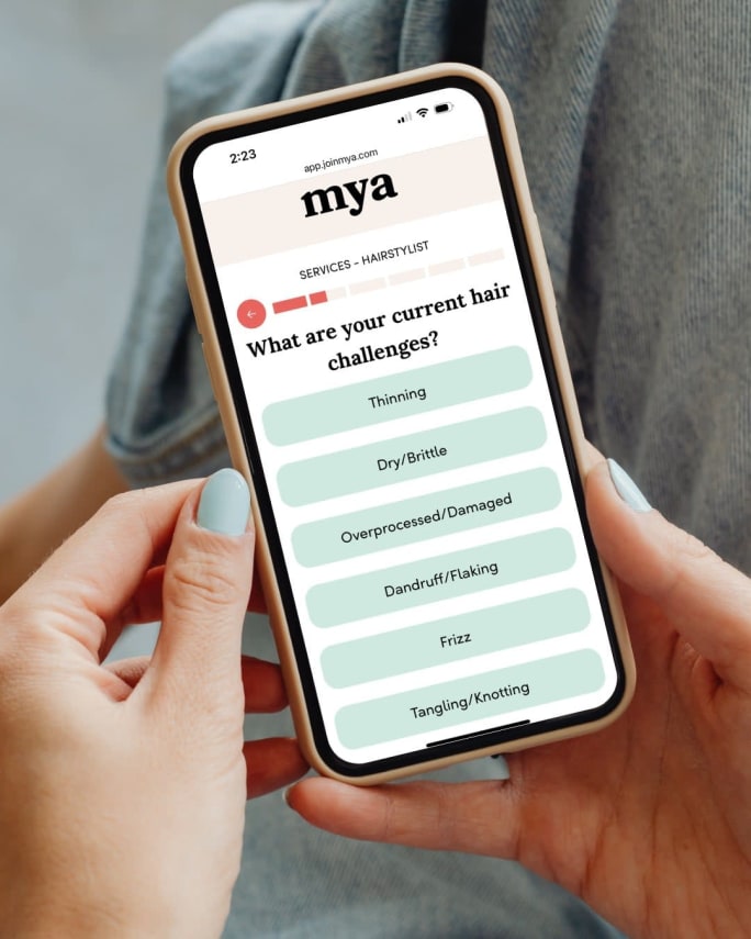 Client holds a smartphone showing a salon matchmaking quiz from Mya that identifies hair concerns, illustrating how technology helps match guests with the right stylist.