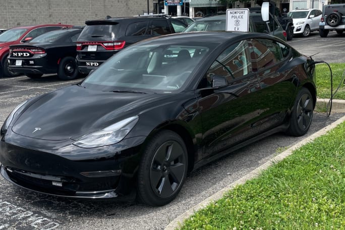 A black unmarked Tesla Model 3 sits in a parking lot, charging.