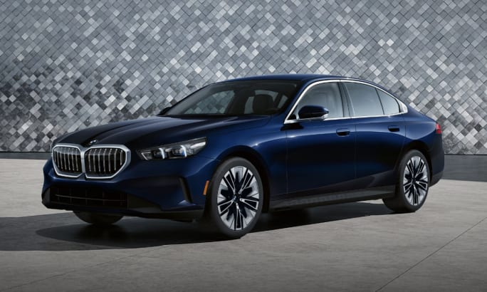 A dark blue 2025 BMW 5 Series Sedan in a gray showroom with a mosaic tiled wall in the background.