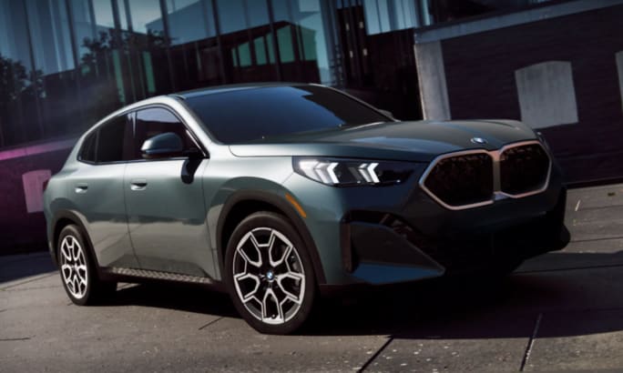 An up close front angle shot of a blue green 2025 BMW X2 in front of a glass building.