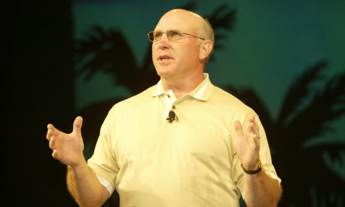 Bill Gibson, in a tan collared polo shirt, speaks on stage.