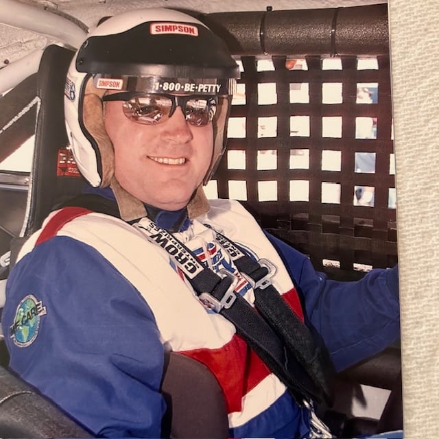 Bill Gibson in the driver's seat of a race car, decked out in racing gear and helmet.