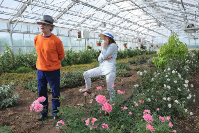 A older man in a greenhouse gazes off camera as his daughter stands behind him.