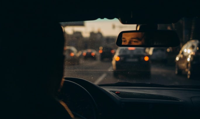 View from the back seat of a car with a man's half-open eyes framed in the rearview mirror. 