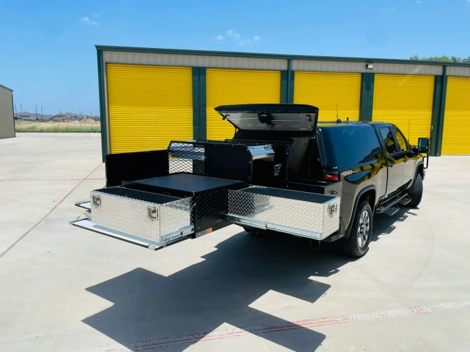Black pickup truck with open diamond-plate storage compartments near yellow storage units