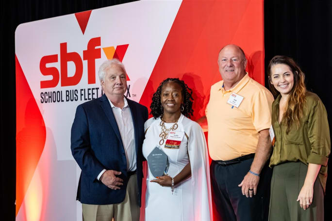  four people stand next to a red and white SBFX banner. in the middle is the award winner in a white dress holding a trophy.