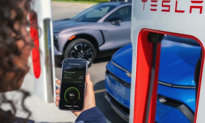 In front of a Tesla charger, a person with curly hair holds a phone out revealing a GM app with charging information.