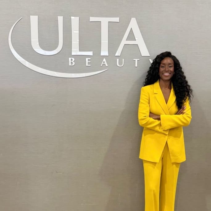 A woman with long black curly hair, wearing a bright yellow pantsuit, stands in front of a wall with an ULTA sign.