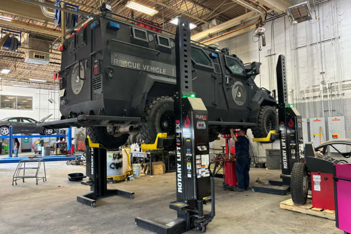 A black armored vehicle receives an oil change while on a vehicle lift.