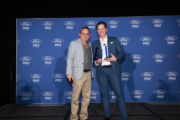 Chris Brown, associate publisher at Bobit/Fleet Group, stands with Matt Atkenson of Ford Pro as Atkenson holds two Commercial Van of the Year awards in front of a Ford Pro backdrop at Work Truck Week.