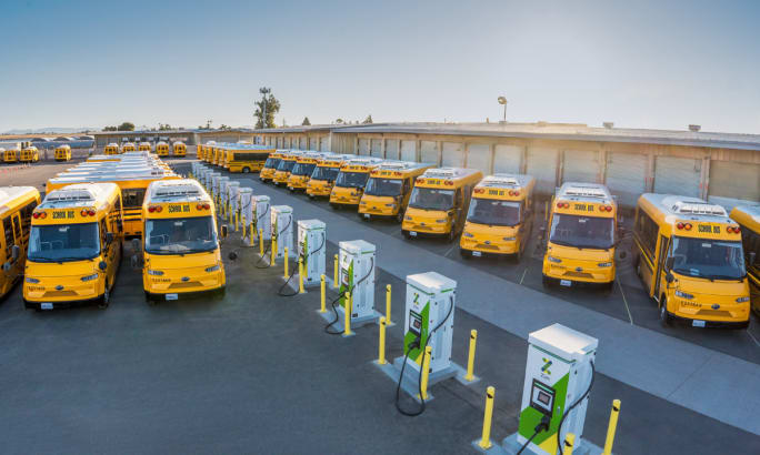 A lineup of electric school buses managed by Zum is shown sitting in a bus lot with Zum-branded chargers.