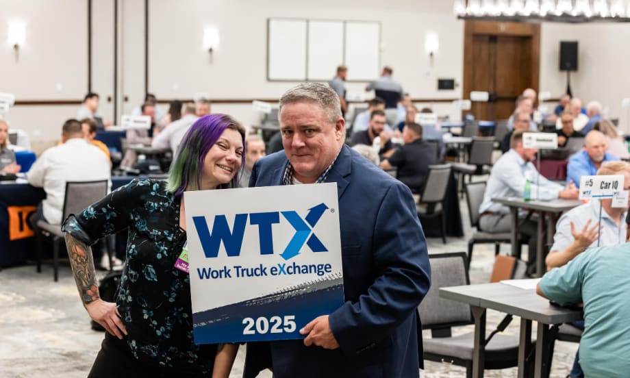 Lauren Fletcher and Wayne Parham pose with a Work Truck Exchange 2025 sign during a networking session while fleet professionals meet in the background.