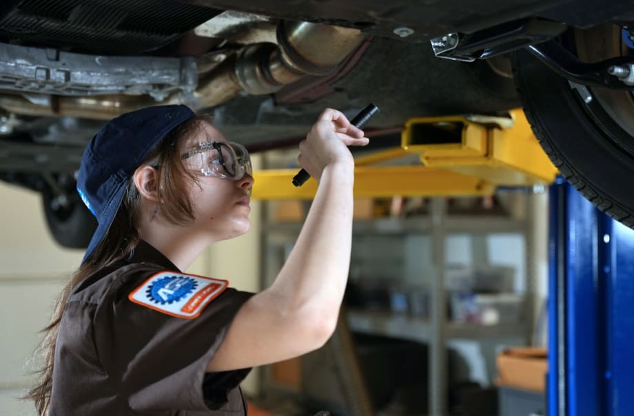 Female automotive mechanic uses a flashlight to check under a vehicle overhead on a lift.