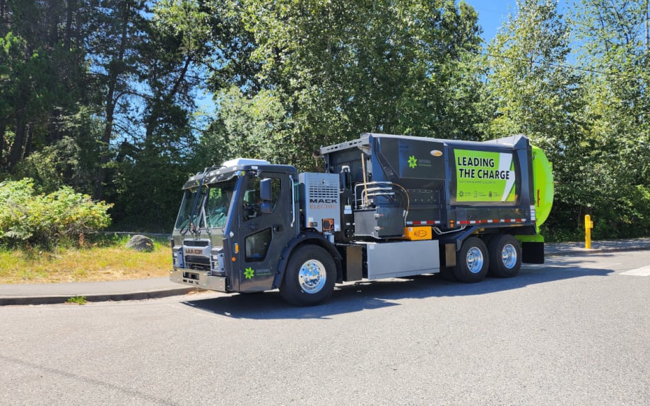 A trash collection truck driving through a street with greenery.
