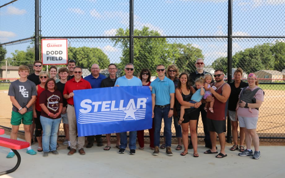 Group photo in front of backstop of a baseball field as people hold a banner that says Stellar.